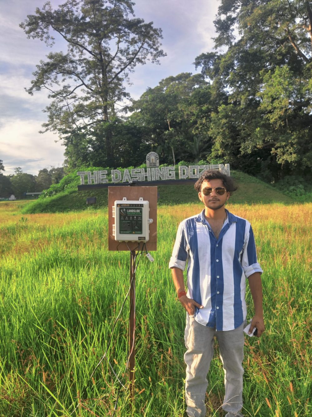 A BHU-Rakshak landslide monitoring unit mounted on a post in a grass field at the pilot site in Assam, with a team member standing beside it.
